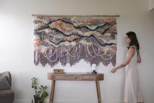 Contemporary woven wall art emphasizing softness and flow, handcrafted with sustainable fiber choices. Shades of purple, pink, ivory, gray, and blue.