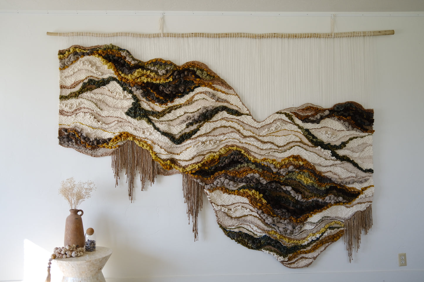 Handwoven wall hanging in earthy neutrals, crafted from wool, cotton, and recycled linen, featuring soft curves and layered texture. Organic shape and fringe.