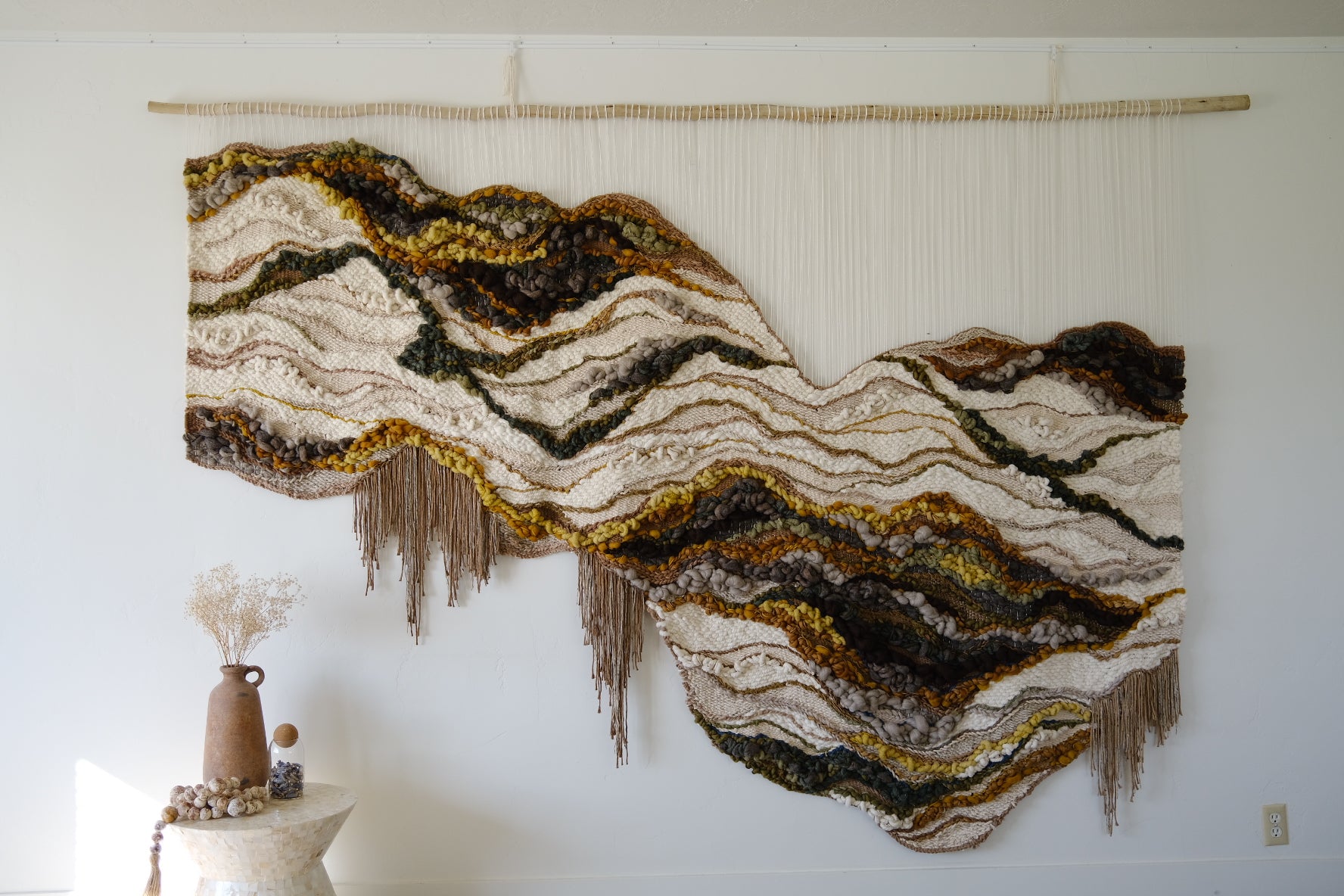 Handwoven wall hanging in earthy neutrals, crafted from wool, cotton, and recycled linen, featuring soft curves and layered texture. Organic shape and fringe.
