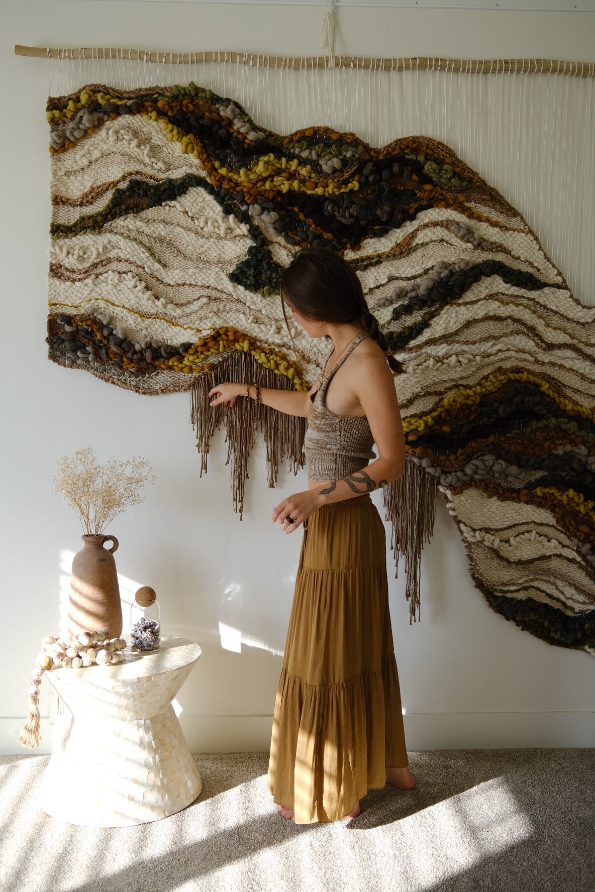 Handwoven wall hanging in earthy neutrals, crafted from wool, cotton, and recycled linen, featuring soft curves and layered texture.  Colors include beige, green, ochre, burnt orange, brown, gray and other earthy tones.