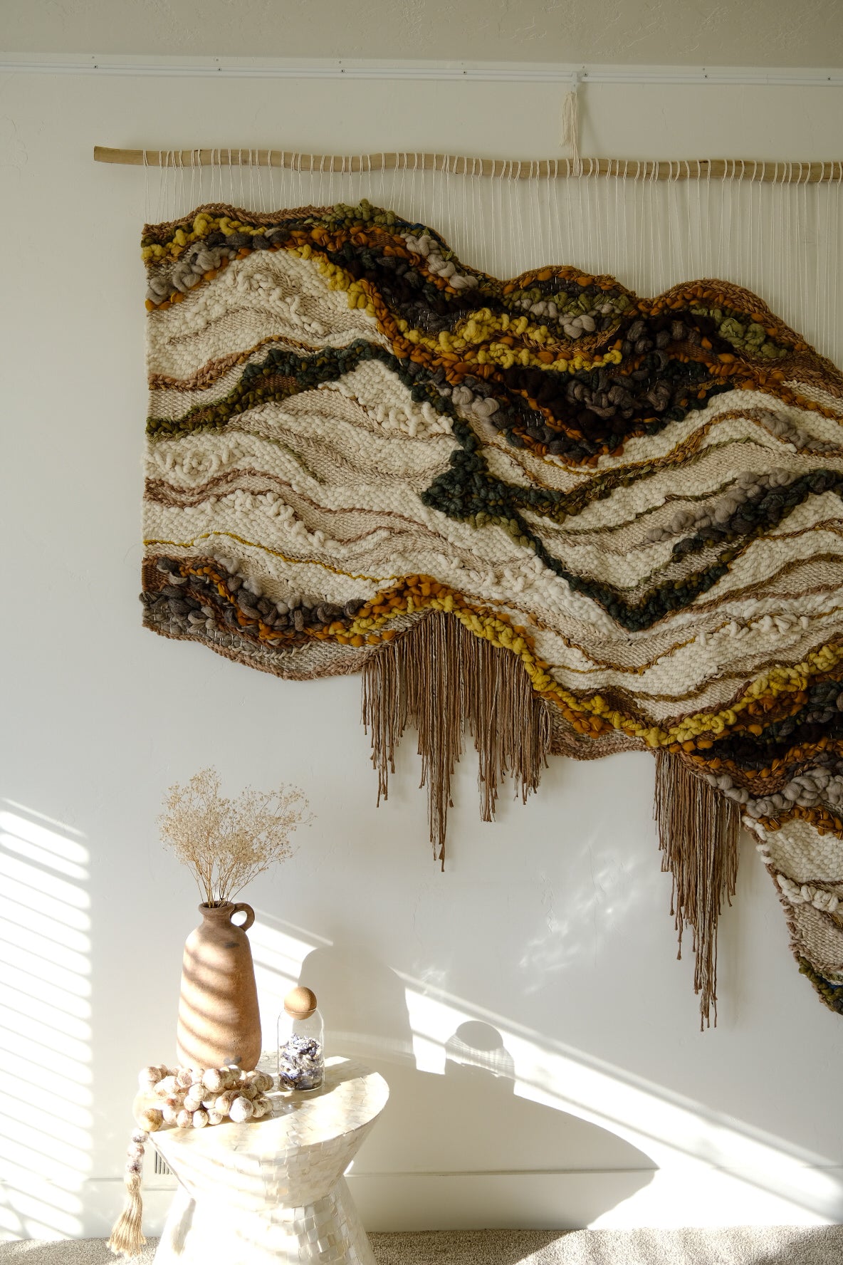 Handwoven wall hanging in earthy neutrals, crafted from wool, cotton, and recycled linen, featuring soft curves and layered texture. Organic fringe and texture.