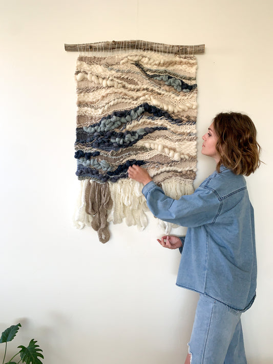 Textural wall hanging inspired by coastal landscapes, featuring undulating patterns in soft blues and neutrals.