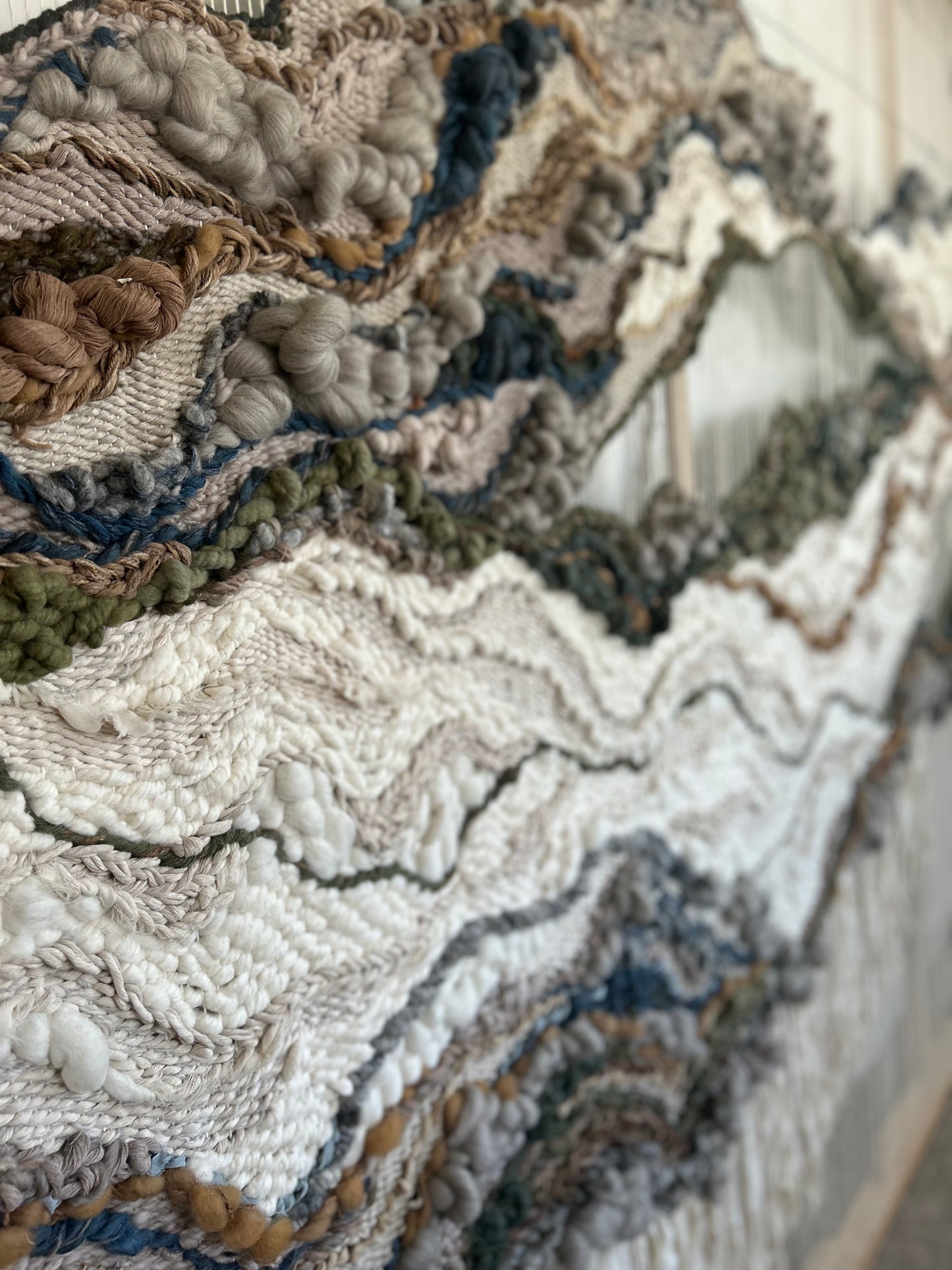 Large-scale woven wall art with undulating forms and natural fibers, designed by textile artist Rebecca Whitaker. Accent colors include blue, green, gray, brown, beige, white, black, and more earth tones.