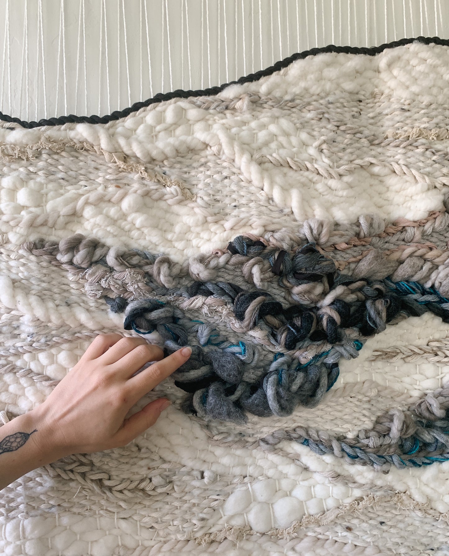 Handwoven fiber art wall hanging featuring intricate textures and organic patterns in natural tones. Accent colors in blue, ivory, beige, gray and other sand tones.