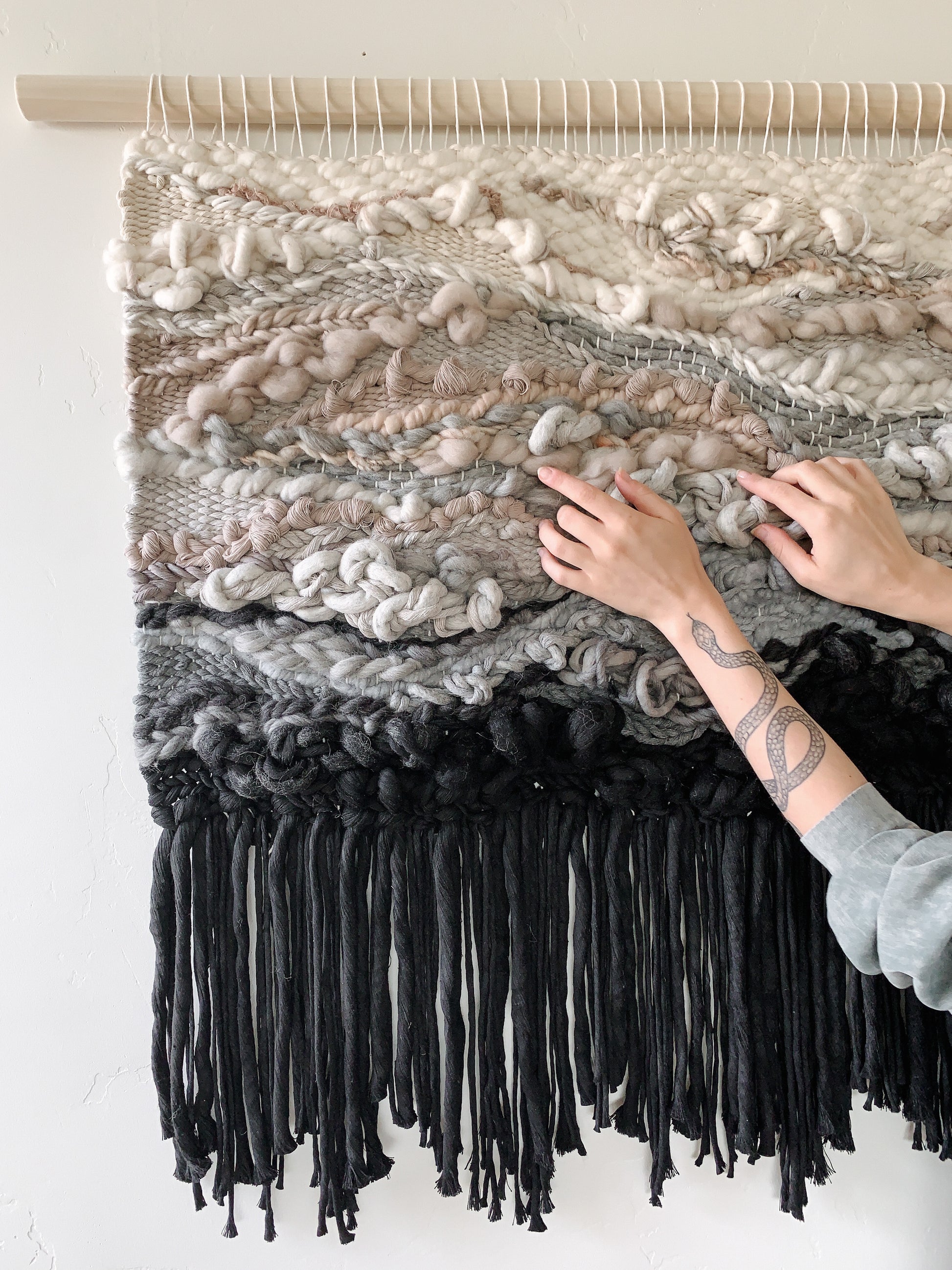 Contemporary woven wall art with layered textures and earthy hues, adding depth to any interior space. Knotted design with heavy fringe. Accent colors in black, gray, sand tones beige, ivory and buff.